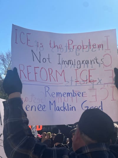 A person holds a sign that reads "ICE is the Problem Not Immigrants." Below this is written "Reform ICE," with the word "ICE" crossed out and "st ICE x." Underneath, it reads "Remember Renee Macklin Good." The image is taken at an angle, showing the person's head and a crowd in the background. The sky is visible in the upper-left corner. The sign is primarily written in black and red.