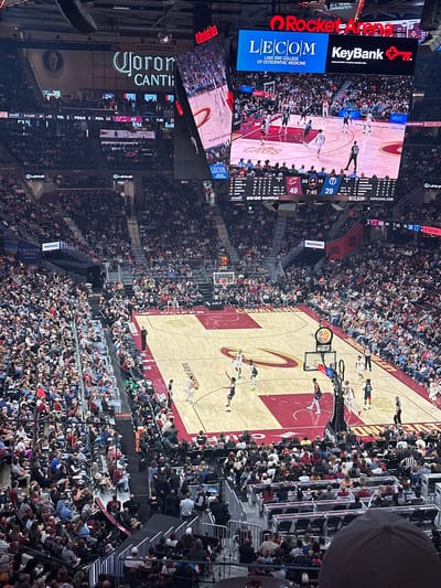 The image shows an interior view of Rocket Mortgage FieldHouse in Cleveland, Ohio, United States of America, during a Cleveland Cavaliers basketball game. The basketball court is visible with players in action, and the stands are densely populated with spectators. A large video screen displays the game footage, statistics, and advertising. Advertisements for LECOM, KeyBank, Corona, Lexus, and other entities are visible. The game score indicates the Cavaliers are leading. The lighting is bright, appropriate for a sporting event. People are generally seated and watching the game. A 50/50 raffle is also being conducted.