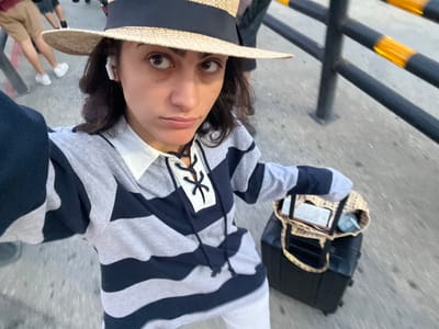In Ferno, Italy, a woman is taking a selfie outdoors. She wears a wide-brimmed straw hat with a black ribbon, an earbud, and a grey and navy striped long-sleeved shirt with a white collar and a laced neckline. She is looking at the camera with a slight pout. To her right, a black suitcase is visible with a leopard-print tote bag on top. The tote bag contains a book and a blue water bottle. The background shows a grey paved ground, a black metal railing with yellow reflective bands, and the legs of other people.