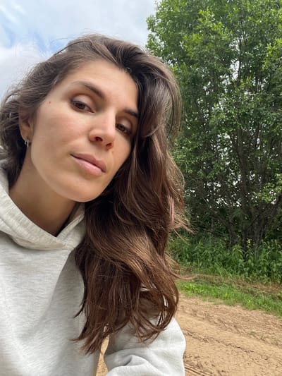 A medium shot shows a woman from the chest up, looking towards the camera. She has long, wavy brown hair that is falling forward, obscuring part of her face. She is wearing a light gray hooded sweatshirt. Her face is tilted slightly, and her eyes are open. She has a mole on her right cheek.
Behind her, the background is an outdoor scene. There is a large green tree with lush leaves, and further in the background, there is green foliage and a dirt path. The sky above is overcast with clouds. The image appears to be taken outdoors, likely in a rural or park setting. The location is identified as Ural'skoe, Russia, though there are no specific visual cues within the image to confirm this location.