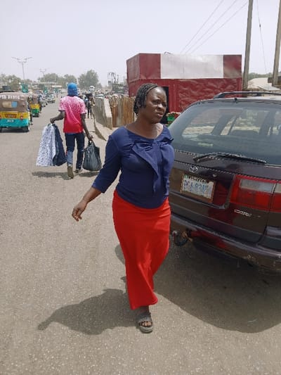 The image depicts an outdoor street scene, described as being in Kadoma, Zimbabwe, under bright, possibly hazy sunlight. In the foreground, an adult female with dark skin and braided hair walks towards the left side of the frame. She wears a dark blue long-sleeved top with a bow-like detail on the front and a long, flowing red skirt, paired with dark sandals. Her head is turned slightly to her right, and she appears to be looking forward. She casts a distinct shadow to her right.

Immediately to her right, a dark-colored Toyota Corolla station wagon is partially visible, parked on the road. Its rear license plate, "BRA 9243," is partially legible. The car has a trailer hitch assembly visible beneath the bumper.

In the midground, behind the woman, a male figure walks away from the camera, carrying several patterned fabric items in his left hand and a dark bag in his right. He wears a red patterned t-shirt, dark trousers, and a blue cap. Further left and behind him, a light blue and yellow three-wheeled vehicle (auto-rickshaw/tuk-tuk) with "PTA 10106" visible on its side is parked or moving slowly. Other vehicles and pedestrians are visible in the distant background, along with streetlights and utility poles.

On the right side of the frame, behind the parked car, a large, rectangular structure, possibly a shipping container, is visible; it is painted red with a large white band across its top. The road surface is asphalt, and the sky in the background appears light and hazy.