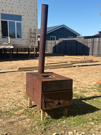 The image depicts an outdoor scene in a rural or semi-rural setting, potentially in the city of Matchino, Russia, characterized by construction and general disarray. In the foreground, a rusty, rectangular metal stove with a tall smokestack stands on makeshift supports. The stove has a small, hinged door on the front and appears to be in a state of disuse. The ground surrounding the stove is a mixture of dirt, sand, and patchy grass, with scattered cigarette butts and a blue plastic object visible.
Behind the stove, a construction site is evident. A partially completed building, clad in white insulation panels with black circular fasteners, is to the left, featuring a white-framed window. A wooden ladder leans against the building. Further back, wooden planks and construction materials are stacked on the ground. A wooden fence encloses the area, with a dark-colored house visible beyond it. The overall impression is one of ongoing construction or renovation in a somewhat untidy outdoor environment. The sky is clear and blue, indicating sunny weather.