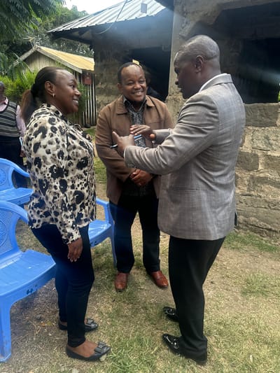 The image depicts an outdoor gathering of at least four adults, likely in Kiganjo, Kenya, under natural daylight. The overall mood appears friendly and engaging, with several individuals smiling.

In the foreground, a woman dressed in a black and cream leopard-print blouse and black pants is seated on a blue plastic chair, looking up and smiling. Beside her, a man in a brown jacket and a dark patterned shirt is also seated, actively engaging in conversation. He smiles broadly and appears to be holding a small, light-colored object in his hands, possibly a phone or a notebook. He wears brown dress shoes.

Towering over the seated individuals, a man in a light grey patterned suit jacket and black dress pants stands, leaning forward as he converses with the seated man. He has a shaven head and wears shiny black dress shoes. A fourth person, wearing a striped shirt, is partially visible in the lower-left corner.

The setting is an open-air space with green grass underfoot. The background features rustic architecture, including a stone wall and a corrugated metal roof structure, possibly part of a building or shelter. Lush green foliage and trees indicate a natural, possibly rural or semi-urban environment. The bright, natural lighting suggests it is daytime, and the positive expressions on the faces of the visible subjects convey a sense of enjoyment and interaction. No text is visible in the image.