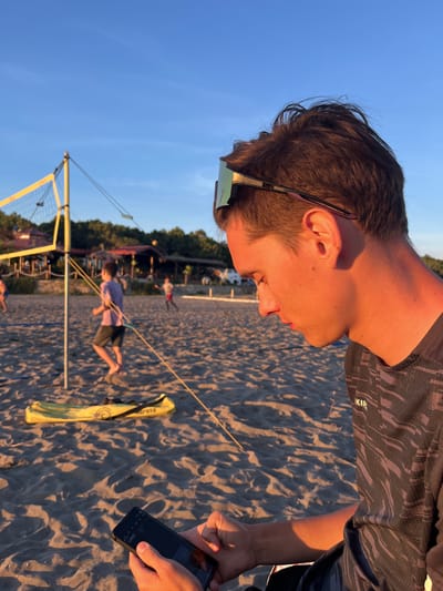 A young Caucasian male is seated on a sandy beach in front of a volleyball net. He is wearing sunglasses on his head and a dark, patterned t-shirt. In his hands, he is holding a black smartphone, looking down at it. The beach is illuminated by the golden light of a sunset. In the background, there are other people playing volleyball, a volleyball net, and trees. The scene is set in Pistula, Montenegro, as indicated by the context.