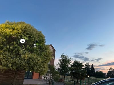The image depicts an outdoor scene at dusk, with a clear blue sky transitioning to soft hues of pink and orange near the horizon. The location is identified as Ponteterra, Italy.
In the foreground, a large tree with dense, variegated green and yellow foliage is prominently featured on the left side of the frame. Two circular, white lights are attached to the tree, casting a bright glow. The tree is situated next to a brick building, with a portion of its facade visible. The building has orange and blue elements, and a dark glass door. Paved ground is visible in front of the building.
To the right of the tree, the landscape opens up to reveal more vegetation, including smaller trees and a palm tree. Several low-level, spherical lights are positioned along a pathway or fence line in the middle ground, illuminating the area. In the background, a treeline is visible against the colorful sky, with hints of structures or rooftops peeking through. A car is partially visible in the lower right corner of the image.
The overall atmosphere is serene and twilight-like, suggesting a calm evening in an urban or suburban setting in Ponteterra, Italy. There are no people or animals visible in the image. The focus is on the architectural elements, the natural environment, and the artificial lighting.