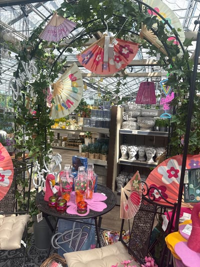 The image shows an outdoor market stall in Fritzlar, Germany, featuring a variety of decorative items, primarily fans and garden ornaments. The stall is adorned with ivy and other greenery, creating a somewhat whimsical atmosphere. In the foreground, a small round table displays several pink and orange plastic cups with straws, a pink flamingo figurine, and small glass candle holders. Behind the table, a dark metal chair is partially visible, decorated with a large pink fan featuring a floral pattern.
Further back, shelves are stocked with decorative pots and bust-like ornaments. A neon sign spelling "Cocktail Bar" is visible in the background, suggesting a festive or celebratory theme. Several other fans, in various sizes and colors, are hung from an arched structure covered in greenery. The overall impression is of a brightly lit, summery display of goods. The location is identified as Fritzlar, Germany, though the specific context of the market or the exact stall within the city is not provided. No people are visible in the image.