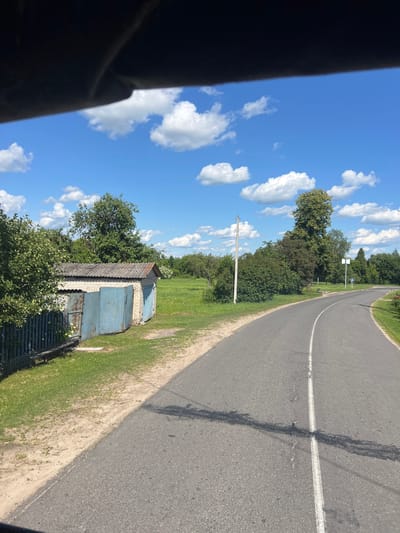 This image depicts an outdoor scene on a sunny day. The sky is a vibrant blue with scattered white, puffy clouds. A paved road curves to the right, bordered by a grassy verge. Adjacent to the verge, there are several small, weathered brick buildings with blue metal doors, possibly garages or storage sheds, partially obscured by lush green trees and foliage. A simple wooden fence runs in front of some of the structures. In the background, more trees create a natural backdrop. The lighting suggests it is daytime, likely mid-morning or afternoon, with bright sunlight casting shadows. There are no people visible in the image. The overall impression is a rural or semi-rural setting in Belarus, characterized by a peaceful, sunlit atmosphere.