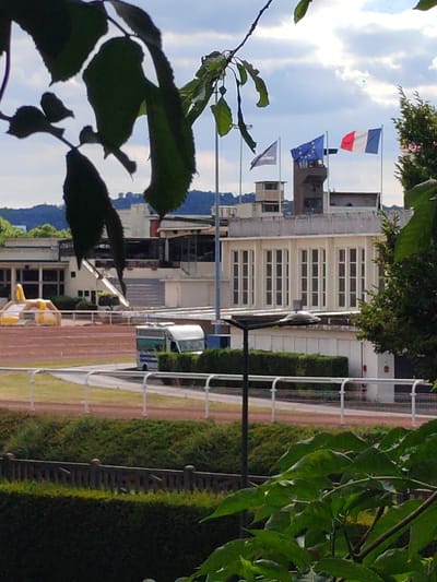 The image depicts a scenic view of a racecourse, likely in Soisy-sous-Montmorency, France, on a partly cloudy day. In the foreground, dark green leaves partially obscure the view, creating a natural framing effect. Beyond the leaves, the racecourse itself is visible, with a reddish-brown track bordered by white railings. A white bus is parked near the track, and further back, there's a yellow inflatable structure.
Dominating the mid-ground is a modern building with a distinctive facade featuring rows of tall, rectangular windows. Several flags are prominently displayed on poles in front of this building. From left to right, these appear to be a triangular flag, the European Union flag, and the French flag. A control tower-like structure is also visible behind these flags.
The background shows a hilly landscape covered in trees, suggesting a semi-rural or suburban setting. The sky is a mix of blue and white clouds, indicating moderate weather conditions. The overall impression is one of a calm, possibly early afternoon scene at a sporting venue. No people are clearly visible, and there is no discernible text in the image.