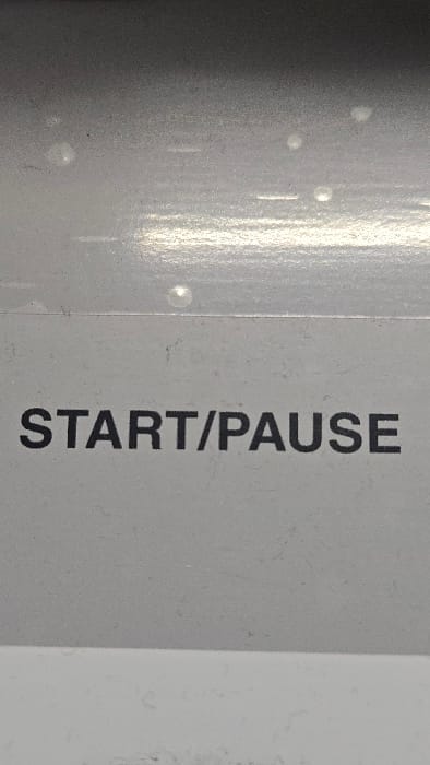 The image depicts a close-up, vertical view of a light gray control panel surface in Lompoc, United States. The text "START/PAUSE" is rendered in black, sans-serif uppercase letters, centered horizontally in the lower portion of the frame. The surface exhibits a fine texture with visible dust particles and several scattered, irregular circular light reflections, predominantly in the upper half. A faint horizontal line or seam runs across the frame approximately one-third from the top, separating a slightly darker gray area above from the lighter gray section containing the text below. No people, active subjects, or specific interactions are present.