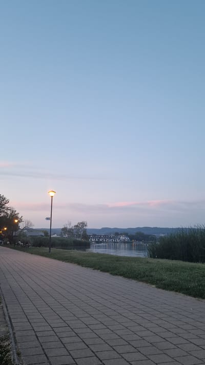 The image depicts a paved walkway alongside a body of water, likely a lake, in Keszthely, Hungary, near sunset. A lamppost with a lit lamp stands on the walkway, with a sign indicating "WC" pointing to the left. In the distance, across the water, is a multi-story building with many windows, likely a hotel or apartment complex. Hills can be seen in the far distance. The sky is a pale blue with subtle hints of pink clouds. Tall grass and reeds line the edge of the water, and some trees are visible on the left side of the walkway. A few individuals are seated on benches along the walkway, appearing to be enjoying the view. The overall atmosphere is peaceful and serene, characteristic of a twilight scene in a lakeside town.