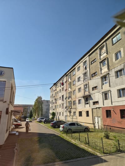 The image depicts a residential street scene in Fălticeni, Romania, characterized by multi-story apartment buildings and parked cars under a clear blue sky. The foreground shows a paved area with interlocking pavers, transitioning to a walkway and a street.
On the left side of the image is a light-colored building with a prominent dark sign featuring a stylized blue 'Ñ' logo. A person is walking away from the camera on the walkway, carrying a white plastic bag. Further down the street, a white van with orange and red markings is parked.
The main focus of the image is a long, multi-story apartment building dominating the right side of the frame. This building, with its concrete facade, exhibits a somewhat weathered appearance. Several windows are visible, some with laundry hanging on clotheslines. Balconies and external brackets are also present.
Across the street from the apartment building, another similar apartment building is visible in the background, set further back. Adjacent to the main apartment building on the right, there is a section with a reddish-brown facade. A small, fenced-off grassy area with a young tree is situated in front of this section of the building.
Several cars are parked along the street, including a silver hatchback, a dark sedan, and a white SUV. The street itself is asphalt. The overall environment suggests a typical urban residential area.