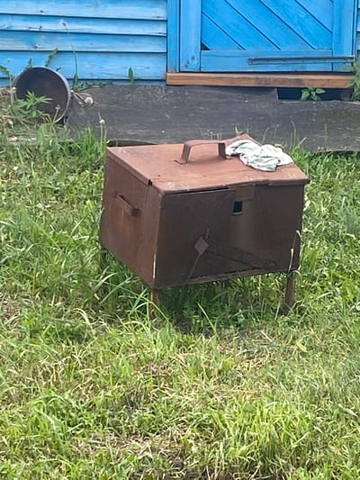 In Urazovo, Russia, a weathered, rusty metal box sits on four thin legs in a grassy area. The box has a hinged lid with a handle and a small square opening on its side. A piece of white cloth with green and blue stripes rests on top of the lid. Behind the box, a weathered wooden structure painted bright blue with horizontal planks is visible, suggesting it is part of a building. Green grass and some weeds surround the metal box, indicating an outdoor, possibly neglected, setting.