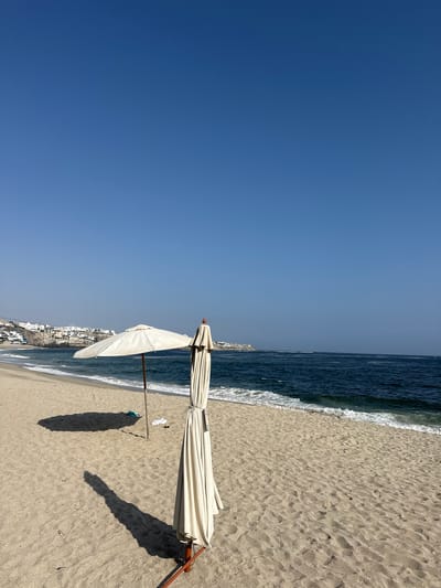 This image depicts a beach scene in Punta Hermosa, Peru, under a clear blue sky. The right side of the image shows a sandy beach with a partially opened, folded white beach umbrella casting a shadow on the sand. The sea is visible along the upper-right edge, with gentle waves meeting the shore. In the distance, along the coastline, are clustered white buildings, likely part of the town. The left side of the image is dominated by a clear blue sky.
