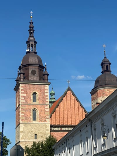 The image shows a view of the towers of St. Margaret's Basilica in Nowy Sącz, Poland, against a clear blue sky. The main tower, made of red brick and stone, is on the left. It features arched windows and a dark, domed belfry topped with a spire and a golden cross. To the right, another smaller tower, also with a dark dome and spire, is visible. A section of the basilica's red brick roof with decorative stonework is also in the frame. In the foreground, a white building with arched windows and decorative trim lines the street, casting shadows from the bright sunlight. The scene is framed by a power line running horizontally across the upper part of the image and a blue pole on the left edge. Trees are visible at the base of the main tower.