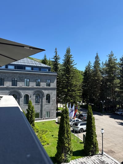 The image captures a view from a balcony or elevated position looking out over a modern building and a surrounding natural landscape. The building, constructed of dark gray stone blocks, features multiple windows and arched entrances. A large, dark gray cantilevered awning extends from the left side of the frame, partially obscuring the upper portion of the building.
In front of the building, a manicured green lawn is visible, bordered by neatly trimmed bushes. Several tall, slender evergreen trees are strategically placed on the lawn, with more dense evergreen forest visible in the background. A paved area, likely a parking lot, is located to the right of the building, with several cars parked there. Among the cars is a white SUV with visible text on its rear door.
In the foreground, to the left, a gray railing is prominent, suggesting it is part of the viewing platform. A lamppost with a white globe light stands near the edge of the paved area on the right. Several flags, including the flag of Armenia, are planted on poles to the left of the paved area, near the lawn. The sky above is a clear, bright blue, indicating a sunny day. The overall setting suggests a resort or a large establishment in a natural, possibly mountainous, environment, consistent with the location of Jermuk, Armenia, known for its spa resorts and surrounding natural beauty.