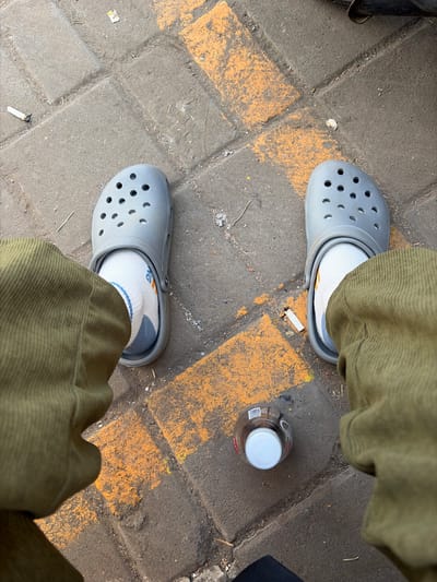 A person wearing grey Crocs and olive green corduroy pants is sitting on a paved surface marked with yellow lines. A bottle with a white cap is on the ground next to the person's right foot. Two cigarette butts are visible on the ground near the person's left foot. The scene appears to be outdoors, possibly in a public area within Secang, Indonesia, given the presence of paved walkways and common urban debris like cigarette butts.