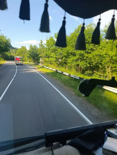 The image shows a view from the inside of a vehicle traveling on a road in a rural, mountainous area. The road is asphalt with white lane markings. On the right side of the road, there is a metal guardrail and a grassy embankment with lush green trees. A statue of a deer stands on the embankment. In the distance, a red truck is visible on the road, possibly a car carrier. The sky is blue with some scattered clouds, suggesting it is daytime. The sun appears to be shining, illuminating the trees and the road. There are decorative dark tassels hanging from the top of the vehicle's interior, partially obscuring the view of the sky and trees. The overall impression is one of a scenic drive through a natural landscape. The location is Ijevan, Armenia. No people are clearly visible.
