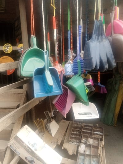 The image displays a retail stall within the city of Kofar Ruwa, Nigeria, featuring a dense arrangement of household cleaning and hardware supplies. Numerous plastic dustpans and hand brushes, in shades of green, light blue, dark blue/black, pink, and white, are suspended by strings in the upper and mid-portions of the frame. Some dustpans contain their corresponding hand brushes. Behind these, various long-handled brooms or mops, exhibiting red, green, blue, and floral patterns, are also hanging, with some individually plastic-wrapped. On the lower left, a partially visible white cardboard box for a "Cut Flex Professional Angle Grinder" rests on wooden planks. Adjacent to it are thin, yellowish rods and a stack of metallic square items. Further right, a multi-compartment wooden container is filled with assorted small hardware fasteners, including nails and screws. A white plastic dustpan is visible on the wooden surface beneath the hanging items. In the background on the left, two circular abrasive cutting discs, one yellow and one orange, are discernible, both labeled "POWER EXTRA." The scene is illuminated by natural light, and no individuals are present.