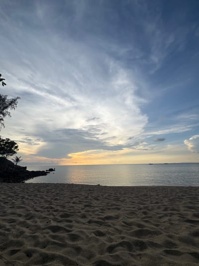 The image depicts a beach scene in Baan Wang Ta Kien, Thailand, oriented vertically. The sky occupies a significant portion of the frame, showcasing a mix of light blue and cloud formations, transitioning to hues of orange and yellow towards the horizon. The horizon line is relatively straight, separating the sky from the sea. The sea appears calm with a reflective surface mirroring the colors of the sky. A strip of sand occupies the right side of the image. Some vegetation is visible in the lower portion of the image along with rocks bordering the beach.