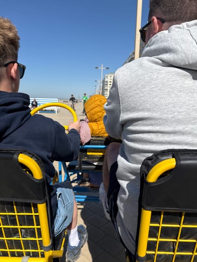 The image depicts an outdoor scene featuring several individuals riding pedal karts on a clear, sunny day. The perspective is from within one of the karts, looking forward and slightly to the right.
In the foreground, to the left, is the back of an adult's head, wearing a light gray hooded sweatshirt. Sunglasses are perched on their right ear. To the right, the back of another person, possibly a child, is visible, wearing a dark blue sweatshirt and holding onto a yellow steering bar of a pedal kart.
The pedal karts are characterized by their bright yellow frames and black padded seats or supports. A black mesh grille is visible on the side of one kart. On a blue panel attached to one of the karts, the word "BINARO" is visible in white text.
Further into the scene, the lower half of another rider is visible, wearing blue jeans and gray athletic shoes with white socks. In the background, other blurry figures of people suggest a lively recreational area, likely a paved promenade or boardwalk. Distant buildings are visible against a bright, clear blue sky, indicating pleasant daytime weather. The scene suggests a leisurely activity taking place in a coastal setting, consistent with Middelkerke, Belgium.