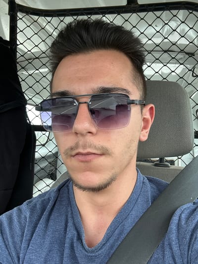 A man is seated in what appears to be the cab of a work vehicle. He is wearing a blue shirt, sunglasses, and a seatbelt. The interior of the vehicle is visible, including the seat and a wire mesh partition behind him. The location cannot be determined to be in Novi Pazar, Bulgaria, from the image alone.