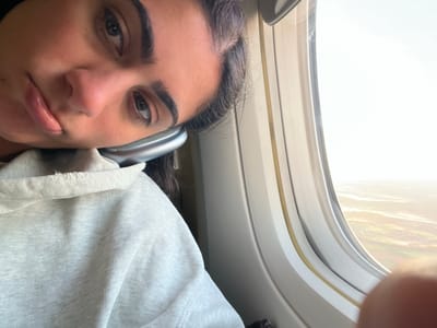 A close-up selfie of a person with dark hair, brown eyes, and a slightly parted mouth, potentially female, taken inside a plane near the window, with the window frame and a blurred view of the outside visible. The person is wearing headphones. The lighting is bright. The location, Meaux, France, cannot be determined from this image alone.