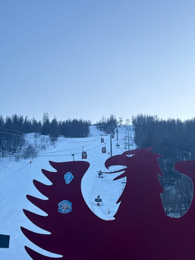 The scene depicts a winter ski area in Saint-Chaffrey, France, under a clear, light blue sky. A large, dark red silhouette of an eagle or phoenix is prominently positioned in the immediate foreground, facing right and partially obscuring the lower and right-hand portions of the landscape. Two circular stickers are affixed to the visible left wing of this silhouette: an upper blue sticker featuring a stylized monkey face with a hat, and a lower multi-colored sticker displaying a crest-like design. Behind the foreground element, a wide, snow-covered ski slope descends from a forested mountain. A gondola lift system, consisting of multiple red enclosed cabins, ascends the slope from left to right along cables strung between support towers. Further to the right and higher on the mountain, a chairlift system with numerous towers and cables is also visible. Dense coniferous forests with dark green trees flank both sides of the ski slope and cover the upper mountain areas. No human figures are identifiable within the visible portions of the lifts or on the slope.