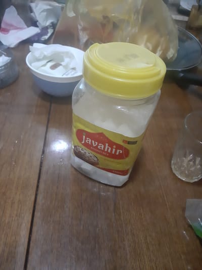 A clear jar of basmati rice with a yellow lid sits on a wooden table. The jar has a label that reads "Javahir" and "BASMATI RICE". A crumpled white tissue is in a blue and white bowl to the left of the jar. A glass is to the right of the jar. The image appears to be taken indoors in a residential setting. There is no indication of location in Sabunchu, Azerbaijan.
