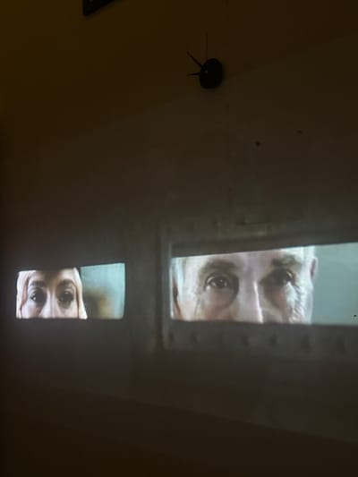 The image shows a projection of two pairs of eyes on a wall. The left projection displays the eyes and forehead of a person with light-colored hair and fair skin. The right projection shows the eyes, nose, and forehead of an older person with graying hair and wrinkles. Both projections are within rectangular frames, suggesting they are displayed on screens or windows. In the upper portion of the image, a black, star-shaped object, possibly part of a clock or decoration, is visible against the dark wall. The environment appears to be dimly lit, with the projections being the primary source of light. The location in Abiansemal, Indonesia, is not visually indicated in the image itself.