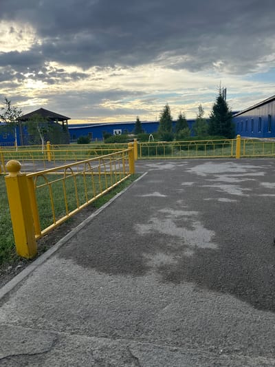 The image depicts an outdoor scene at what appears to be an industrial or educational facility. In the foreground, a yellow metal fence with rounded arches and spherical finials runs along a paved walkway. The walkway is asphalt, with large, irregular patches of lighter gray that suggest water stains or weathering. To the left of the fence is a strip of green grass. In the middle ground, more of the yellow fence encloses a grassy area. Beyond the fence, a dark-roofed gazebo or pavilion is visible, with a birch tree and other greenery nearby. A long, low-rise building with a bright blue facade and numerous windows stretches across the background. Several evergreen trees are planted in front of this building. The sky above is dramatic, filled with dark, heavy clouds interspersed with areas of bright sunlight breaking through, creating a moody atmosphere. The overall impression is of a functional, somewhat utilitarian environment under a dynamic sky. The provided location of Bozshakol, Kazakhstan, suggests this may be a facility within that region.