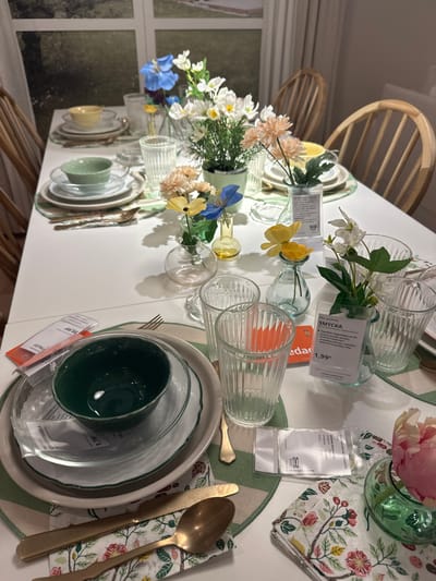 The image shows a beautifully set dining table, likely in a home or a retail display. The table is adorned with floral arrangements in small glass vases, featuring white, yellow, and blue artificial flowers. There are place settings for at least two people, each with a stack of plates, a green bowl, cutlery, and a glass. The cutlery appears to be made of brass, and the napkins have a floral pattern. Several IKEA product tags are visible on the table, one of which mentions "SMYCKA" and "1,99€," indicating artificial flowers. Another tag shows a price of "39,99€." The overall aesthetic is bright and cheerful, with a focus on natural elements and a touch of elegance. The presence of IKEA tags suggests this scene is likely in an IKEA store or a setting inspired by IKEA's design aesthetic. The image does not contain any specific visual cues that would place it in Siero, Spain.