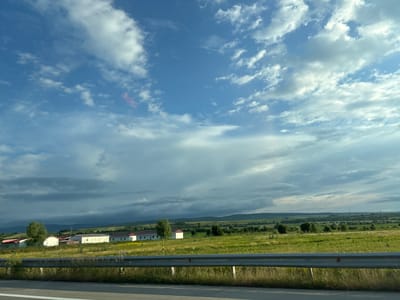 This image depicts a rural landscape with a row of white buildings with red roofs in the foreground, situated in a green field. A metal guardrail runs horizontally across the lower portion of the image, separating the field from a paved road surface. The sky is filled with a mix of blue sky and white, scattered clouds, with some darker clouds present on the horizon. In the distance, rolling hills are visible. The overall scene appears to be a scenic view of the countryside, possibly taken from a vehicle. The location is identified as being in the city of Surami, Georgia.