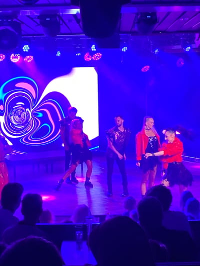 The image depicts a performance on a stage illuminated by vibrant purple and blue lights. The stage features a large, abstract, swirling graphic on a screen behind the performers. Four individuals are visible on stage, dressed in elaborate costumes. Two appear to be dancers, one in a black dress and the other in a black skirt and red jacket. Another individual, a male, is in the background wearing a dark outfit. The audience, partially visible in the foreground, is seated in a dimly lit environment, suggesting a concert venue or theater. The overall atmosphere is energetic and theatrical. This scene is likely taking place at the Hitis, Finland public space, a venue known for hosting performances and events.