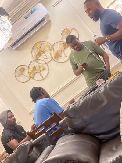 This image captures an indoor gathering of at least five men in what appears to be a modern living room or lounge area, located in Adenta Municipality, Ghana. The photograph is oriented sideways, with the subjects appearing rotated.
On the left side of the frame, two men are visible. One, with a beard, wears a dark blue polo shirt and a gold watch, holding a clear plastic water bottle. Next to him, another man in an olive green t-shirt is looking down, holding a mobile phone and a yellow pen or stylus. In the background behind them, a light-colored wall is adorned with several artistic circular metal wall decorations featuring golden ginkgo leaf motifs. A white Hisense split air conditioner unit is mounted on the wall below these decorations, and part of a decorative light fixture or chandelier is visible at the bottom left.
To the right, large, dark brown or black leather sofas and chairs occupy a significant portion of the frame. Two more men are seated or leaning in this area. One is wearing a blue shirt and has dreadlocks, while another, partially visible at the bottom right, wears a black t-shirt and a grey cap. The overall atmosphere seems casual, suggesting a relaxed social gathering or a meeting among acquaintances. The lighting appears to be artificial, typical of an indoor setting. No strong emotions are discernible from the individuals.