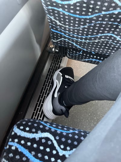 The image shows a close-up view of a person's legs and feet on what appears to be public transportation, possibly a bus or train, in Maardu, Estonia. The person is wearing dark-colored leggings and a black and white athletic shoe with a thick white sole. The shoe has a visible logo that appears to be "Puma". The seating in the vehicle has a distinctive pattern of small gray dots and wavy blue and light blue lines on a black background. A metal grate, possibly part of a ventilation system, is visible on the floor between the seats. The overall lighting suggests an indoor environment, and the composition is from a high angle looking down.