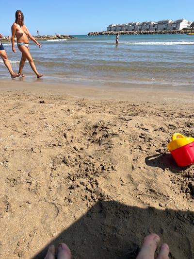 The image depicts a beach scene on a sunny day with a clear blue sky. In the foreground, the sandy beach is visible with footprints and a red and yellow toy bucket. In the mid-ground, a woman is walking along the shoreline, wearing a bikini bottom and with her upper body exposed. Her hair is shoulder-length and brown. Another person's legs are visible to the left of the woman. The sea is calm with gentle waves reaching the shore. In the background, a line of white buildings with blue roofs is visible along the coast, with a breakwater made of rocks in front of them. Further out, a small child is standing in the water. This scene is located at Playa de Alcossebre, within the municipality of Alcalà de Xivert, Spain.