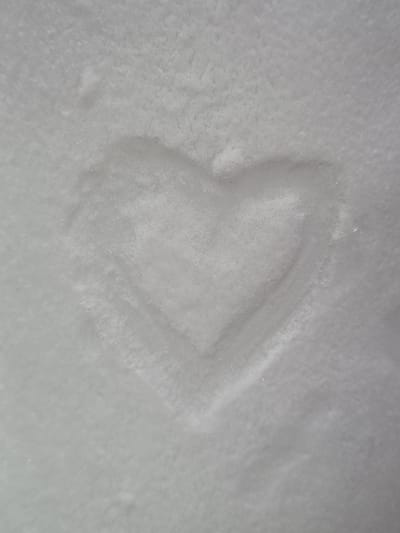 The image presents a direct overhead view of a flat, monochrome surface of compacted snow, located in the city of Kemul, Russia. A distinct heart-shaped impression, approximately centered within the frame and measuring roughly one-third of its overall dimensions, is visible on the surface. The impression creates a shallow, darker depression compared to the surrounding snow, indicative of varied compaction and subtle shadowing. The snow surface exhibits a fine granular and slightly uneven texture, with minute variations in reflectivity. The overall coloration of the scene is a range of light grey to off-white, consistent with snow under diffuse lighting conditions. No individuals, flora, fauna, or man-made objects other than the heart impression itself are present within the frame.
