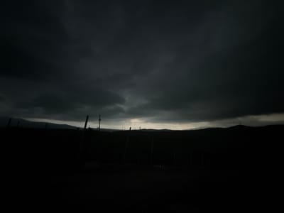 The image depicts a dark, ominous sky filled with heavy, storm clouds, casting a dramatic gloom over a rural landscape. In the foreground, silhouetted against the faint light filtering through the clouds, are several wooden posts, suggesting a fence or some form of agricultural structure. Beyond this, a dark, undulating terrain stretches towards a distant mountain range, also shrouded in shadow. The sky is the dominant feature, with its textured, deep grey and black clouds creating a sense of impending weather. The overall impression is one of a twilight or pre-storm atmosphere in a remote, undeveloped area, possibly on the outskirts of Sisian, Armenia, given the mention of that location. There are no visible people or animals.