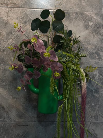 A still life arrangement on a tiled surface featuring a green pitcher or container. The arrangement includes eucalyptus branches with round, dark green leaves; various green leafy plants and stems; and purple-toned floral elements with yellow accents. The tiled surface appears to have a mottled or marbled pattern. The scene is illuminated with moderate lighting.