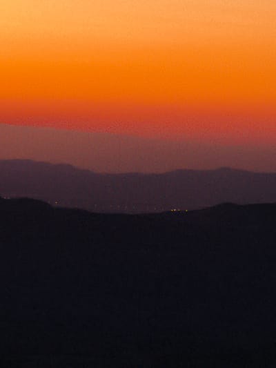 The image captures a sunset over Yeruham, Israel. The sky displays a gradient of warm colors, transitioning from a bright orange at the top to a softer red and then a muted purple towards the horizon. Several layers of distant hills, rendered in shades of purple and dark blue, are visible. The foreground is dominated by a dark, silhouetted landmass, likely a hill or mountain range. Scattered lights, possibly from a town or settlement, are faintly visible on one of the middle layers of hills. The overall impression is one of natural beauty and quietude during twilight.