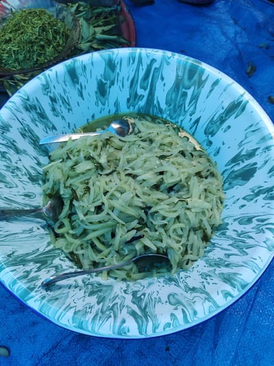 A close-up, high-angle shot shows a large, shallow, blue and white swirled enamel bowl filled with shredded vegetables, possibly papaya or chayote, mixed with green herbs. Three metal spoons are placed within the bowl, partially submerged in the food. The bowl rests on a textured blue tarp. In the upper left corner, a smaller bowl or basket contains a pile of finely chopped green herbs. The background is blurred, but appears to be outdoors with some foliage. The scene suggests a food preparation or serving context, possibly at a market or outdoor gathering in Cintadamai, Indonesia. There are no people visible in the frame.