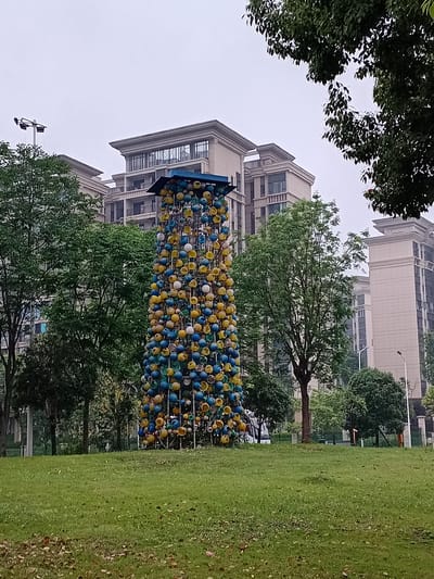 The image captures an outdoor scene in Shuangfu, China, under an overcast sky during the daytime. The central subject is a tall, cylindrical sculptural installation composed of numerous small, spherical or cup-like objects. These objects are primarily colored in vibrant yellow and various shades of blue, creating a distinct mosaic pattern.

The sculpture stands prominently on a well-maintained green lawn, which stretches across the right side of the frame and into the foreground. Scattered green trees with dense foliage are visible around and behind the sculpture, suggesting a park or landscaped green space. In the background, several multi-story residential buildings with a uniform, modern architectural style are present, their light-colored facades contrasting with the gray sky.

No human activity or people are visible, lending a calm and static atmosphere to the scene. Other notable details include the occasional brown leaf scattered on the grass and a street light pole partially visible in the distance near the buildings. The weather appears to be cloudy, indicated by the uniformly pale sky. There is no visible text in the image.