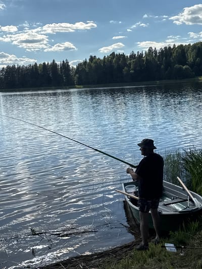 A lone fisherman stands at the edge of a calm lake in Lendži, Latvia, casting a fishing line from a small rowboat. The water reflects the bright blue sky, dotted with fluffy white clouds, and the dense green forest that borders the lake. The fisherman, wearing a hat and shorts, is focused on his activity. The rowboat, containing fishing gear, is partially submerged at the shore. The scene is peaceful and serene, with the natural beauty of the Latvian landscape as a backdrop.