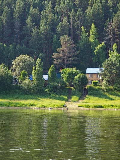 The image depicts a serene lakeside scene with lush green vegetation and a dense forest in the background. In the foreground, the calm water of a lake reflects the surrounding greenery and the bright sky. A narrow strip of grassy shore separates the water from a gentle slope leading up to a cluster of buildings nestled amidst trees. Two wooden houses with blue roofs are visible, one larger and two-storied, and another smaller, single-story structure. A pathway with wooden steps ascends the slope towards the houses. A red flag is planted near the smaller building. On the shore, a small, dark object, possibly a bench or a piece of equipment on wheels, sits near the water's edge. The overall impression is one of a peaceful, rural retreat in a natural setting, likely a dacha or a small settlement along the riverbank in Pristan', Russia.