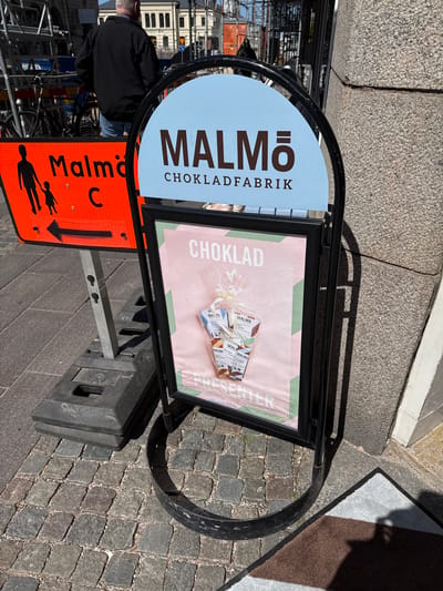 The image shows a freestanding sandwich board advertisement for "Malmö Chokladfabrik", a chocolate factory in Malmö, Sweden. The sign is situated on a cobblestone surface, presumably a sidewalk, next to a street. The A-frame stand, constructed from black metal, supports two panels. One panel, with a light blue background, displays the words "MALMÖ" in large, bold, white letters, and "CHOKLADFABRIK" in smaller, brown letters. The other panel has a light pink backdrop with green stripes near the top. On the panel, there is an advertisement for "Choklad" (Chocolate) and "Presenter" (Gifts) and a collage of Malmö Chokladfabrik chocolate products.
Beneath the sandwich board, there is a weighted black plastic base. Further down, there's an orange traffic sign with a black pedestrian crossing symbol and a black arrow indicating a walking route towards "Malmö C". A bicycle rack is visible behind the sandwich board, along with buildings in the background that could be typical architecture in Malmö. A person dressed in black is visible on the left side of the picture.