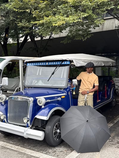 An outdoor scene captures a young man standing beside a blue, vintage-style open-top electric passenger vehicle. The location is in the city of Shinan District, China, indicated by the Chinese characters "奥帆海洋文化旅游区" (Olympic Sailing Marine Culture Tourism Zone) displayed on a banner above the vehicle's windshield.
The man, with dark skin, wears a black flat cap, a light brown short-sleeved polo shirt with white trim, khaki cargo shorts with a white drawstring, and a dark wristwatch on his left wrist. He is positioned on the passenger side of the blue vehicle, leaning his left arm on the support post for the side mirror. His right hand is resting on his shorts. He looks downwards and to his left.
The vehicle is a multi-passenger electric shuttle, designed to resemble a classic automobile, featuring a prominent chrome grille, circular headlights, a chrome front bumper, and silver fender trim. It has a light-colored canvas canopy roof. A small brass-colored bell hangs from the passenger side mirror support. The vehicle is parked on asphalt pavement with white painted lines.
A large, black, open umbrella rests on the ground directly in front of the vehicle's passenger-side front wheel. In the background, green trees with light-colored foliage are visible, along with a grey building facade behind the tree line. A white vehicle, partially obscured, is parked to the left of the blue vehicle.
