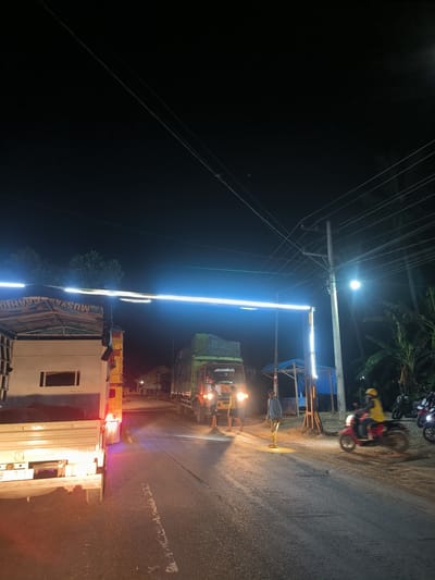 This nighttime scene captures a bustling roadside in Kulu, Indonesia, illuminated by various artificial light sources against a dark sky. The setting appears to be a rural or semi-urban area, with a road leading into the distance.
In the foreground, a white truck with its red taillights glowing is positioned on the left side of the road. Its canvas tarp covering bears partially visible, inverted text that appears to be "MUSYAWARAH." Further down the road, a green truck is parked or slowly moving, with its headlights illuminating the path ahead. On its cab, the word "BISMILLAH" is clearly visible, and its license plate reads "BG 8923."
A prominent feature is a bright, horizontal blue-white light bar spanning across the road, creating an archway effect, possibly indicating a checkpoint, entrance, or temporary structure. To the right of this light bar stands a utility pole laden with power lines, and a street light casting a strong glow. Adjacent to the pole is a small, open-sided structure with a blue tarp roof, possibly a roadside stall or shelter.
Several individuals are present on the road. Near the green truck, two men are standing, one in a light-colored shirt and another in darker attire. On the far right, a person wearing a yellow helmet and jacket is riding a red motorcycle, captured with motion blur, indicating movement. Other motorcycles are parked further right.
The dark sky and reliance on artificial illumination confirm the late hour. Trees and dense foliage, including tropical-looking palm trees, line the right side of the road. The overall scene depicts active nighttime transit and social interaction around a temporary or permanent roadside point.