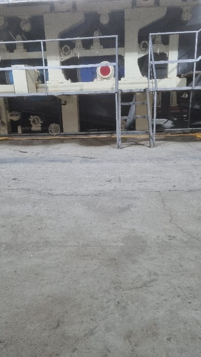 The image shows industrial machinery with a metal framework. Some components are painted white and blue, with a red circular detail. The machinery is positioned next to a gray concrete surface, which dominates the right side of the frame. The image is taken from a slightly elevated perspective.