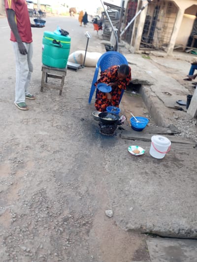 The image depicts a street scene in Shendam, Nigeria, featuring individuals engaged in daily activities. In the foreground, a person wearing a maroon and yellow shirt and light grey trousers stands next to a large, turquoise cooler on a wooden stool. Nearby, another individual, a woman dressed in a colorful patterned dress, is seated on a blue plastic chair, holding a blue plate. She appears to be preparing food, with a cooking pot on a stove and several buckets of water around her. A small plate with food is placed on the ground in front of her. The background shows a street with buildings and other people, suggesting a bustling urban environment. The ground is paved with concrete and dirt, and some debris is visible. The overall impression is one of everyday life and commerce on a Nigerian street.