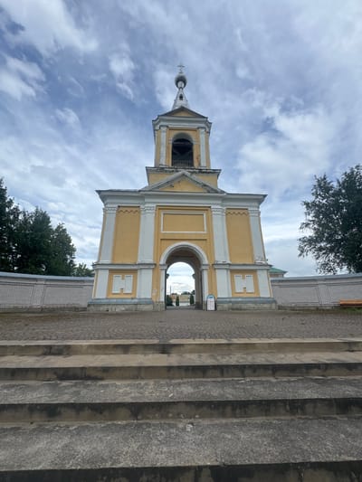 The image shows a yellow and white bell tower in Рассвет, Russia. The bell tower is the central focus of the image and is framed by a stone wall on either side. In front of the bell tower is a paved courtyard with a set of concrete stairs in the foreground. The sky above is cloudy. There are trees on the left and right sides of the bell tower, and in the background, a gate is visible through the archway of the bell tower. There are no people visible in the image.